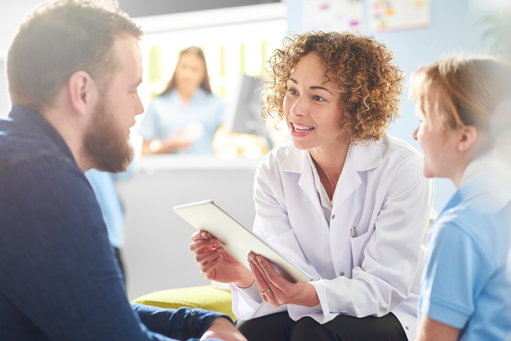 A female pharmacist sits with a father and sick daughter in the pharmacist consultation area and discusses his prescription and choice of medication viewing the details on a digital tablet. In the background a senior woman and is at the counter being served by a female pharmacy assistant .